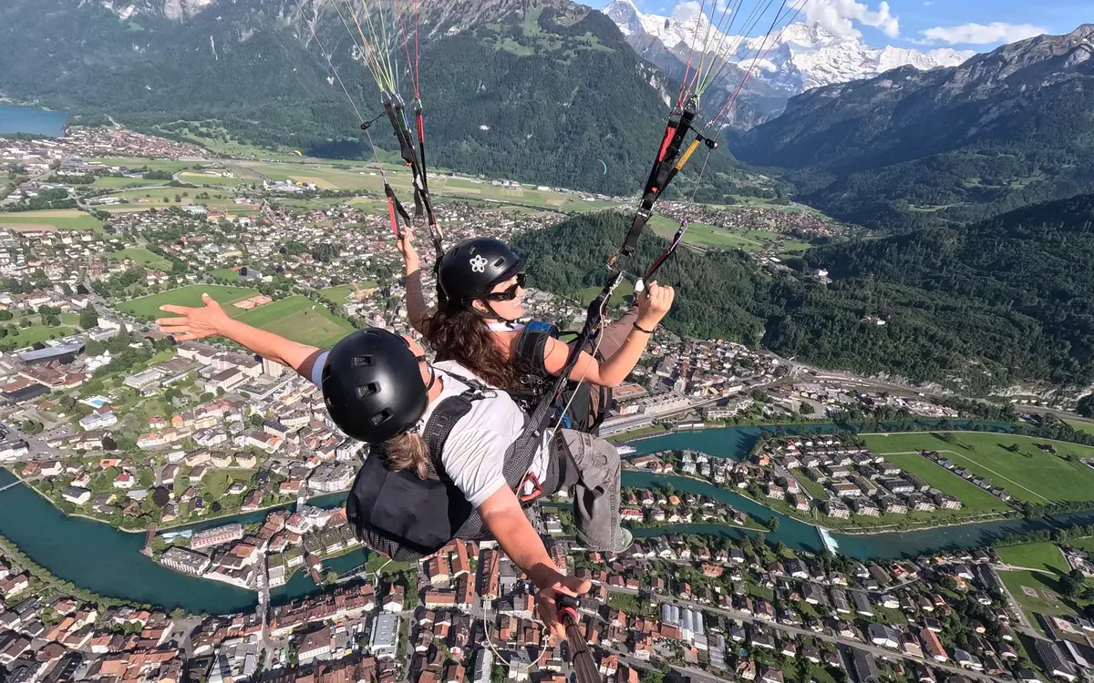 A paraglider descending towards open green meadows with a lake and hills beyond