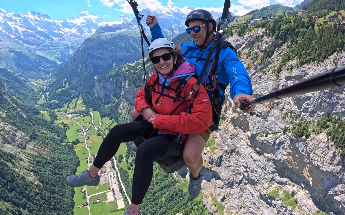 A tandem glider over alpine meadows with a passenger looking down
