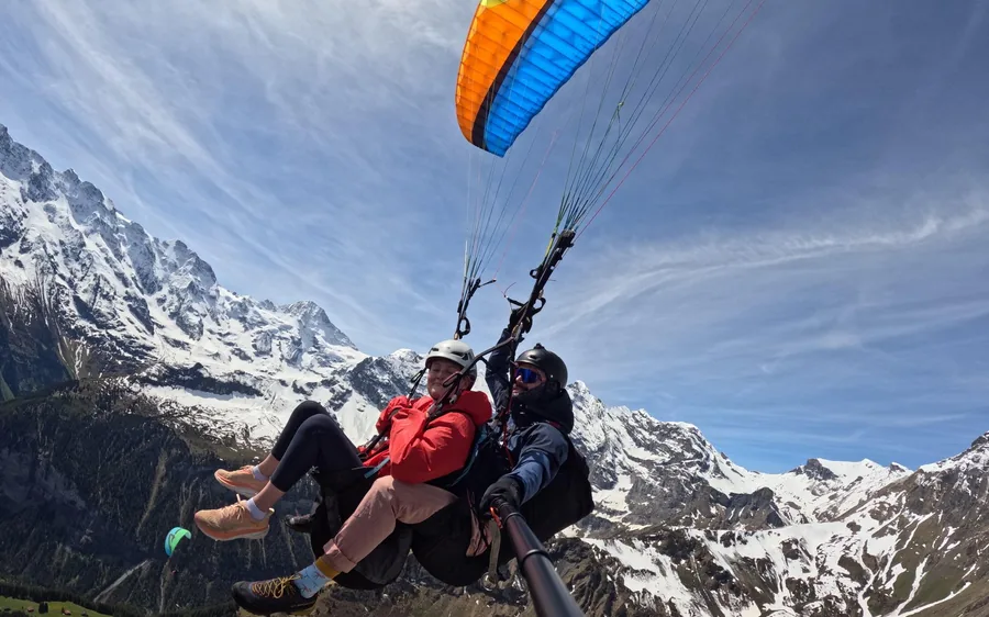 Two people under one paraglider wing over a deep valley with snow peaks behind